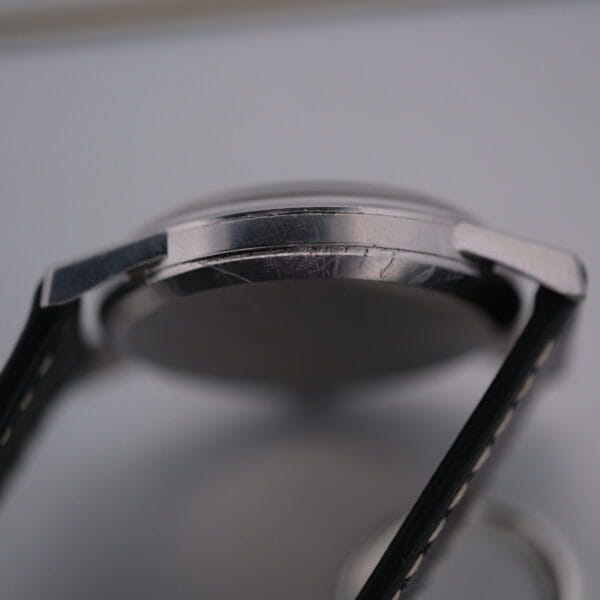 Side close-up of the Omega Manual Wind Cal. 268 wristwatch, highlighting its metallic case and attached black leather strap with scratches adding character. The watch face is intriguingly out of focus.