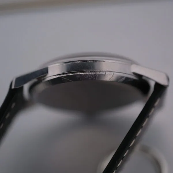 Side close-up of the Omega Manual Wind Cal. 268 wristwatch, highlighting its metallic case and attached black leather strap with scratches adding character. The watch face is intriguingly out of focus.