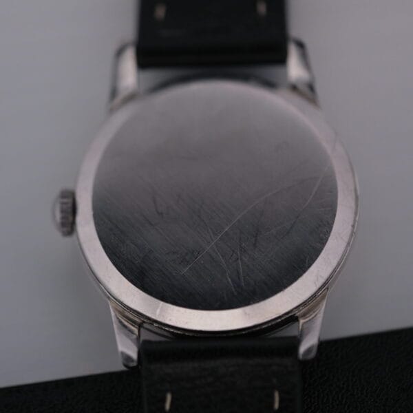 A close-up of the scratched back of an Omega Manual Wind Cal. 268, featuring a round silver design and a black leather strap, placed diagonally on a gray surface.