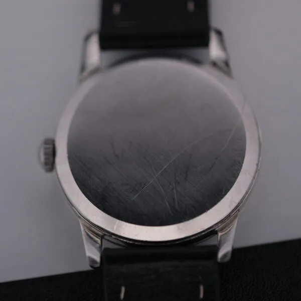 A close-up of the scratched back of an Omega Manual Wind Cal. 268, featuring a round silver design and a black leather strap, placed diagonally on a gray surface.