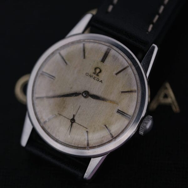 The Omega Manual Wind Cal. 268 features a cream face with bold black hour markers and two sleek hands. It has a black leather strap, elegantly displayed on a dark surface.
