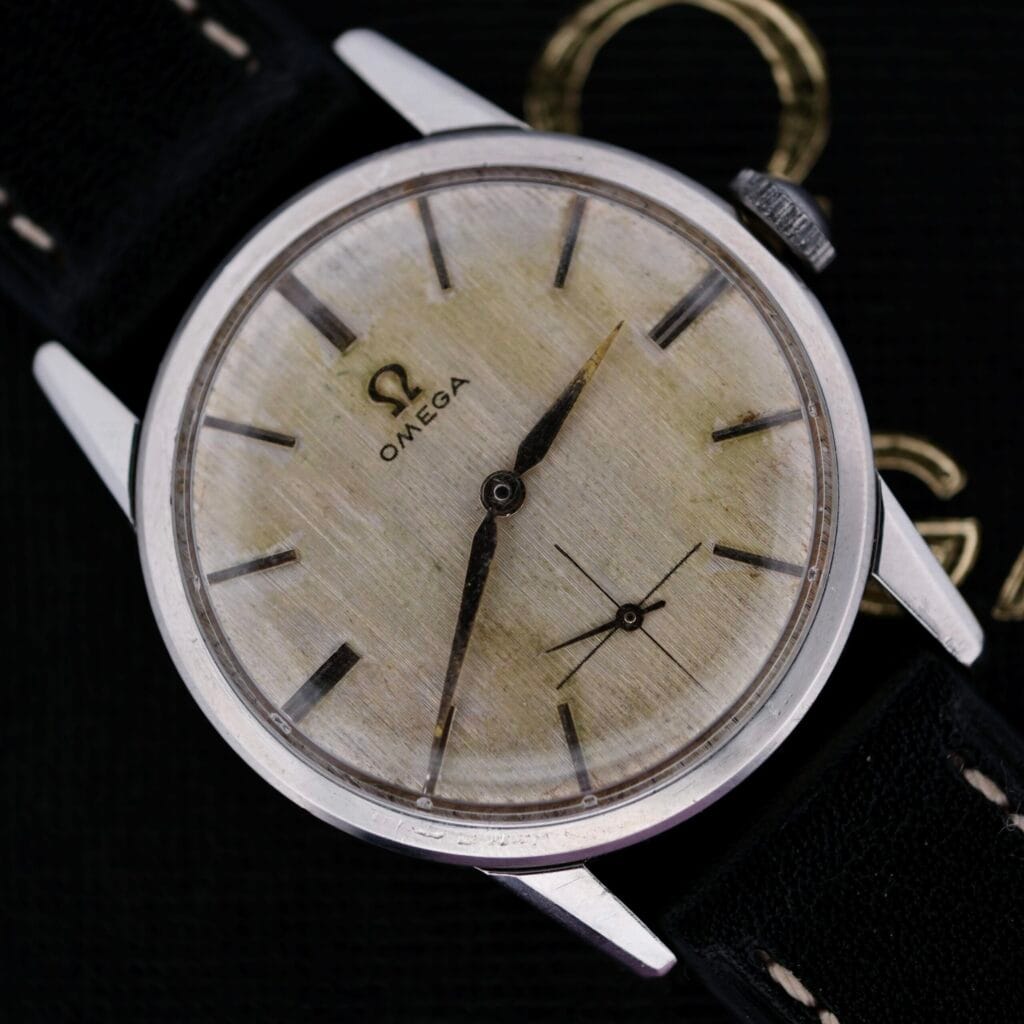 Close-up of a weathered silver dial on an Omega Manual Wind Cal. 268, with black hour and minute hands and a small subdial. It features a black leather strap, dramatically set against a dark background.