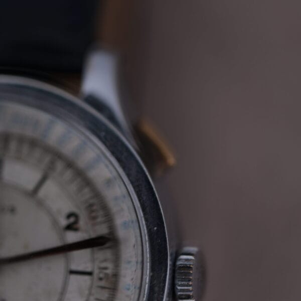Close-up of the Cevrus Monopusher Chronograph Sector Dial, showcasing its vintage look with a worn dial, visible hour and minute hands. The partially shown watch face on the left focuses on the silver case and crown against a blurred brown background.