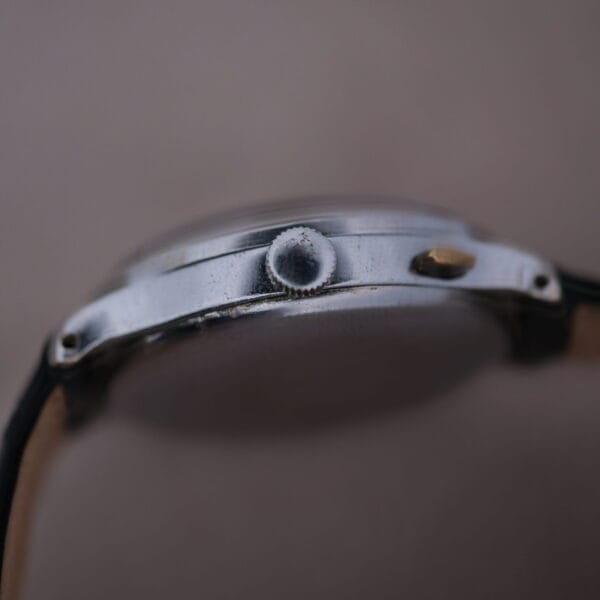 Close-up of the Cevrus Monopusher Chronograph Sector Dial wristwatch, with a round textured crown and slightly tarnished metallic case, complemented by a leather strap. The side profile is highlighted against a soft, blurred background.