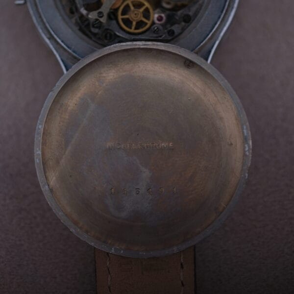 The image reveals the intricate workings of a Cevrus Monopusher Chronograph Sector Dial with its back cover removed, exposing the gears inside. Engravings on the inner cover read nickel chrome and 114491. Set against a soft brown background, it captures timeless craftsmanship.