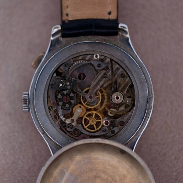 Close-up of a Cevrus Monopusher Chronograph Sector Dial wristwatch with its back cover removed, revealing intricate gears and mechanisms. It features a beige leather strap and silver casing, set against a soft, neutral background.