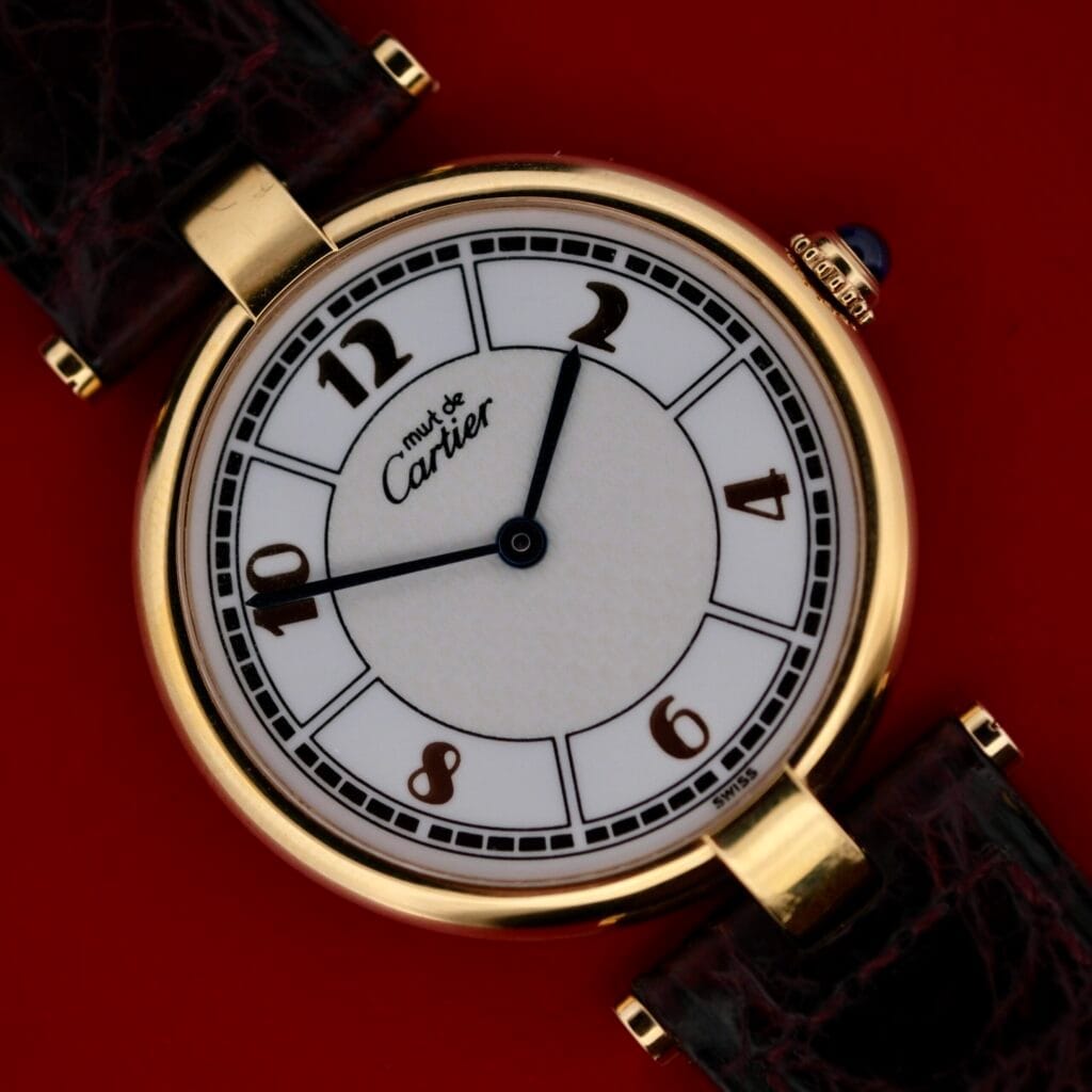 A Cartier Vendome Reference 1861 Vermeil 30mm wristwatch featuring a round gold case, white dial with black numerals, displayed on a bold red background.