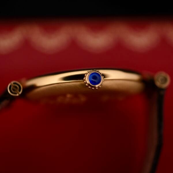DSCF1859 A close-up of the Cartier Vendome Reference 1861 Vermeil 30mm watch crown glistens in gold with a striking blue gemstone on a rich red background. Its case features intricate detailing around the crown, echoing timeless elegance.