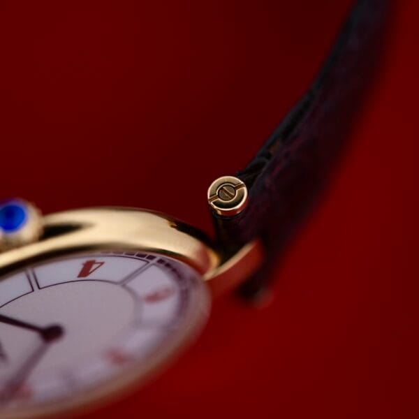 DSCF1861 Close-up of a Cartier Vendome Reference 1861 Vermeil 30mm wristwatch against a red background. It features a white dial with black and red markings, gold casing, and black leather strap. The crown has an engraved logo.