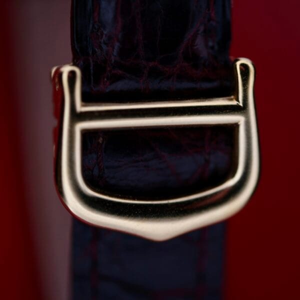 DSCF1862 A striking Cartier Vendome Reference 1861 Vermeil 30mm watch features a shiny gold buckle on a dark leather strap, framed by a rich red background. The buckle showcases a distinctive rectangular shape with curved edges, reminiscent of vintage design.