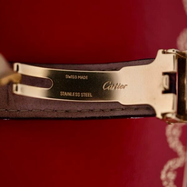 DSCF1863 A close-up of the Cartier Vendome Reference 1861 Vermeil 30mm clasp, engraved with Swiss Made and Cartier Stainless Steel, is attached to a dark brown leather strap, set against a red background with gold detailing.