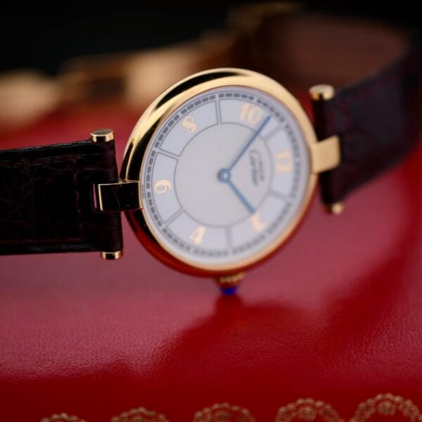 DSCF1866 A close-up of the Cartier Vendome Reference 1861 Vermeil 30mm, a vintage luxury wristwatch with a gold case and dark leather strap. The watch face is simple, featuring gold numbers and hands on a white background, elegantly placed on a rich red leather surface.