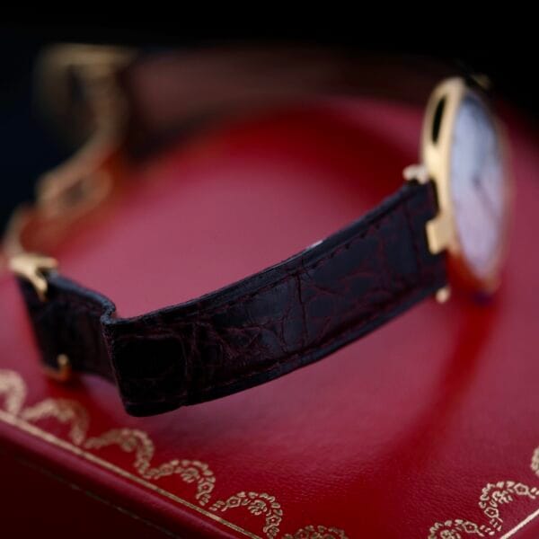 DSCF1867 A close-up of the Cartier Vendome Reference 1861 Vermeil 30mm, featuring a dark leather strap and a gold case, rests on a red ornate-patterned box. The focus is on the strap and case, exuding timeless elegance against a softly dimmed background.