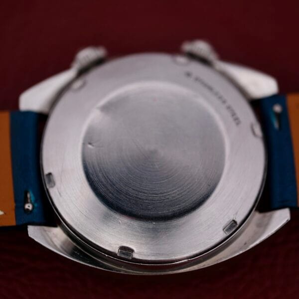 The image displays the back of a silver 1971 Jaeger LeCoultre Memovox Egg wristwatch case on a brown surface. Its blue and brown strap complements the smooth case back, featuring minor marks and the inscription STAINLESS STEEL.