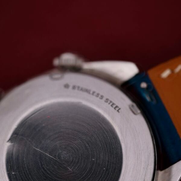 Close-up of the back of a 1971 Jaeger LeCoultre Memovox Egg stainless steel wristwatch, featuring a scratched surface. STAINLESS STEEL is engraved along the top edge. The watch has a brown leather strap with visible stitching, set against a dark maroon background.