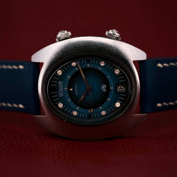 A close-up of a 1971 Jaeger LeCoultre Memovox Egg wristwatch with a blue leather strap on a red background. It features a round blue face, silver casing, dual knobs, date display set to the 12th, and silver hour markers.