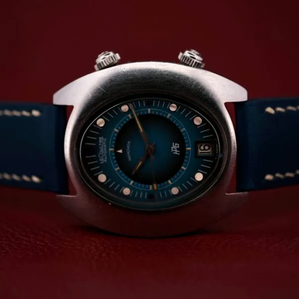 A close-up of a 1971 Jaeger LeCoultre Memovox Egg wristwatch with a blue leather strap on a red background. It features a round blue face, silver casing, dual knobs, date display set to the 12th, and silver hour markers.