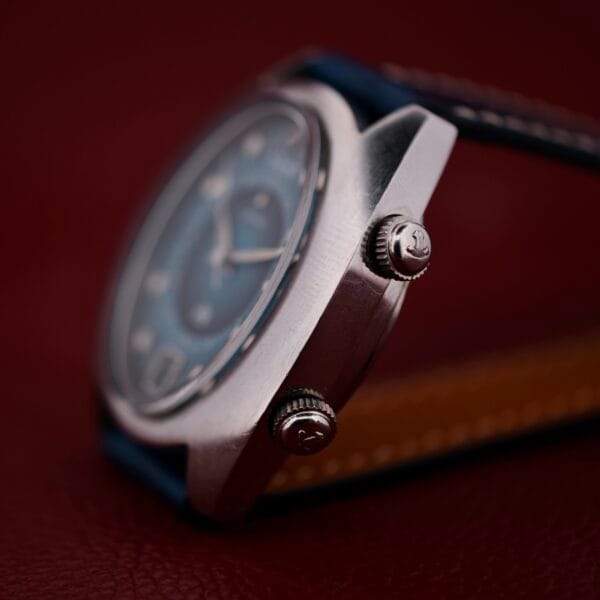 A close-up of the 1971 Jaeger LeCoultre Memovox ’Egg’ wristwatch, showcasing its blue dial and dual crowns on a brown textured surface, with the watch case in focus and the strap and dial softly blurred.