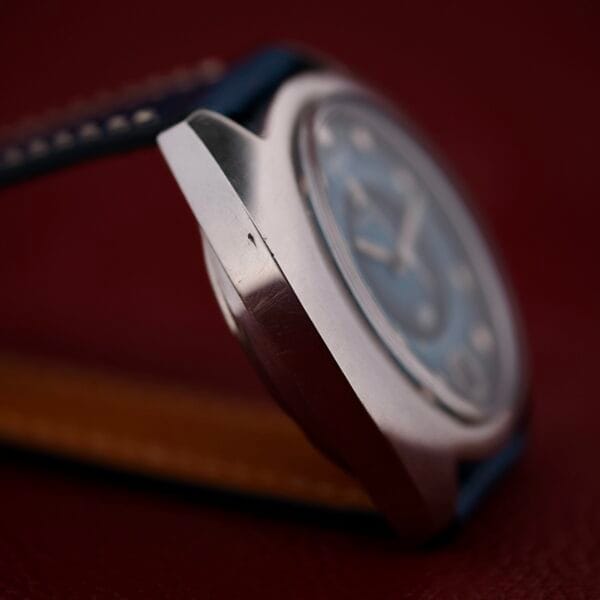 Close-up of a 1971 Jaeger LeCoultre Memovox ’Egg’ with a stainless steel case and blue dial, featuring a blue leather strap with white stitching. The deep red background beautifully highlights the side profile of this elegant watch.