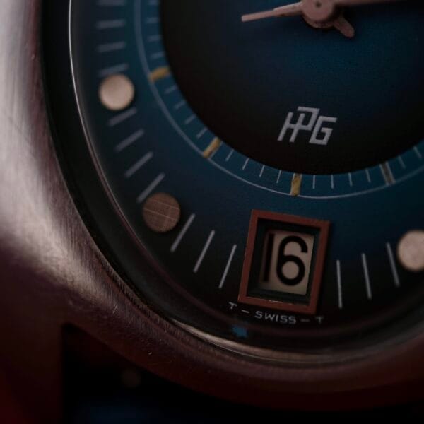 Close-up of a 1971 Jaeger LeCoultre Memovox Egg watch face, showcasing a blue dial with a date window at 16, luminescent hour dots, and a metallic case. The initials HPG and Swiss are elegantly visible on the dial.