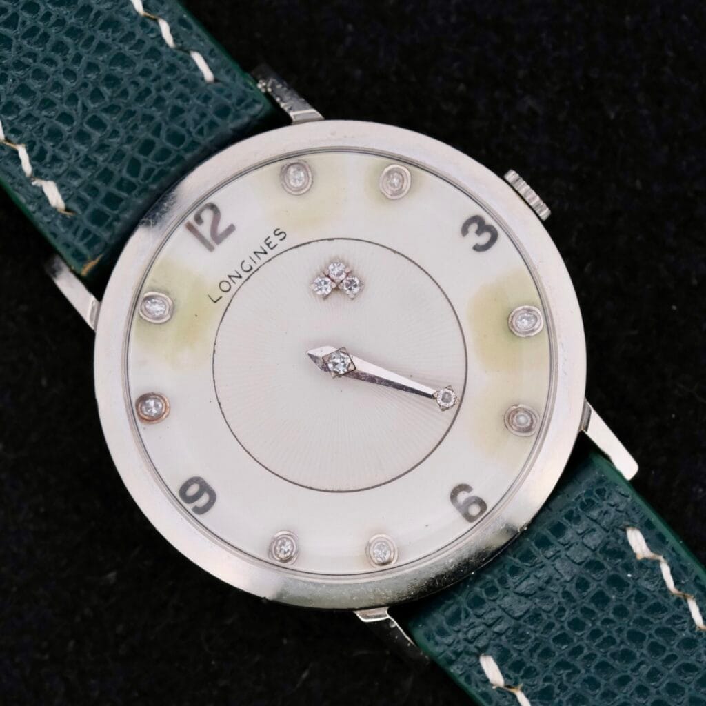 The 1958 Longines Mystery Dial wristwatch, crafted in 14k white gold, features a silver and white face with dark green leather strap. It showcases silver hour markers at three, six, nine, and twelve oclock with two decorative center diamonds.