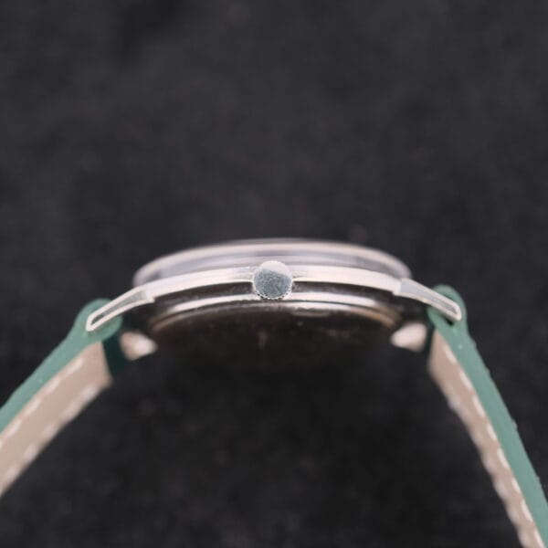Close-up of a 1958 Longines Mystery Dial wristwatch crown, highlighting the green band and metallic casing against a dark background.