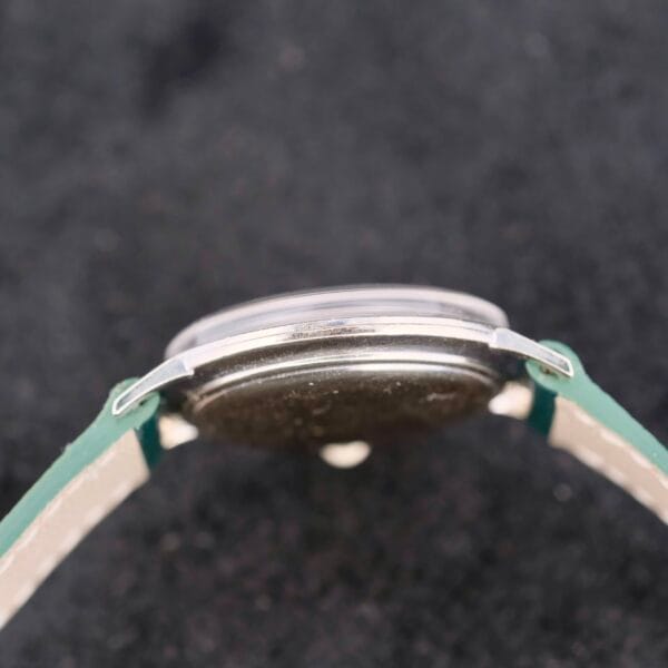 A close-up side view of the 1958 Longines Mystery Dial 14k white gold wristwatch with diamonds, featuring a green strap against a black background, highlighting the sophisticated casing and strap attachment.