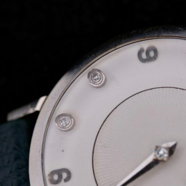 Close-up of a 1958 Longines Mystery Dial wristwatch face with a white dial, featuring silver hour markers at 3, 6, and 9 oclock adorned with round diamonds. A sleek silver hour hand contrasts the textured circular background.
