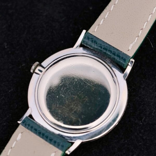 The image showcases the back of the 1958 Longines Mystery Dial watch in 14k white gold with diamonds, featuring a smooth, round case back. It is paired with light leather straps accented by dark green edges and rests on a dark, textured surface for classic contrast.
