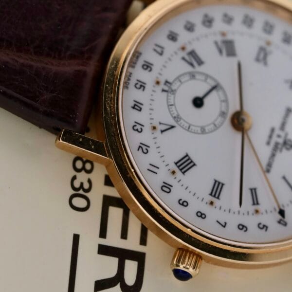 Close-up of a Baume Mercier 18k Gold Calendar wristwatch featuring an analog display, brown leather strap, 18k gold case, white dial with Roman numerals, small sub-dial, and a crown with a blue gemstone. Background has partial black text on white.