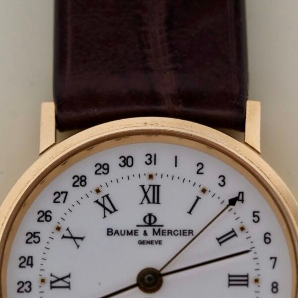 Close-up of a Baume Mercier 18k Gold Calendar watch, Ref 15162, featuring a brown leather strap. The white dial displays Roman hour numerals, Arabic date numerals, and the iconic Phi logo. It boasts an exquisite gold bezel.