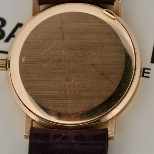 A close-up of a Baume Mercier 18k Gold Calendar watch case, engraved with 181, 523052, and 75162, showcases its elegance. It features a dark brown leather strap against partial black text on a white surface.