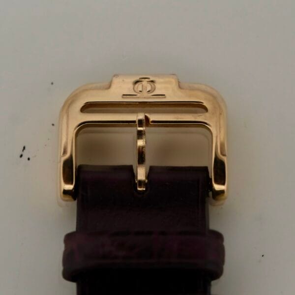 The close-up highlights the Baume Mercier 18k Gold Calendars ornate watch buckle attached to a dark leather strap. Its emblematic design, reminiscent of the Calendar Ref 15162, stands out elegantly against a light backdrop.