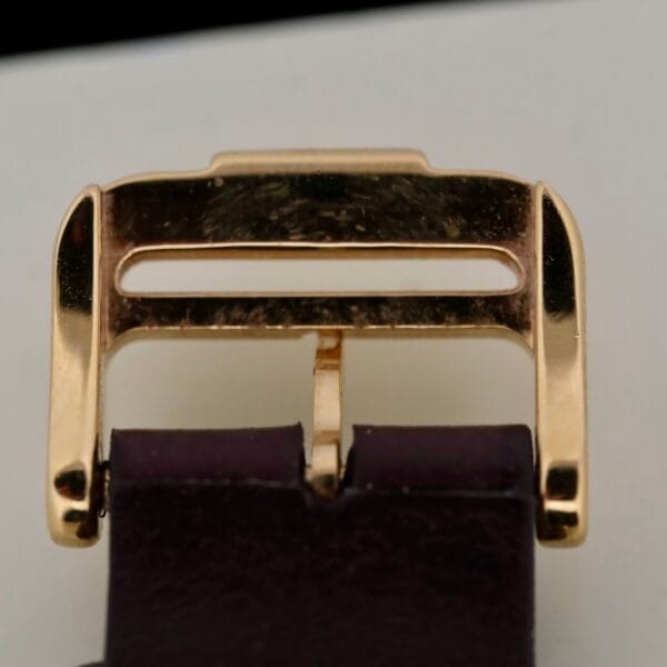 Close-up of the gold-colored Baume Mercier 18k Gold Calendar watch buckle, with two horizontal bars and prong, attached to a dark leather strap on a white background.