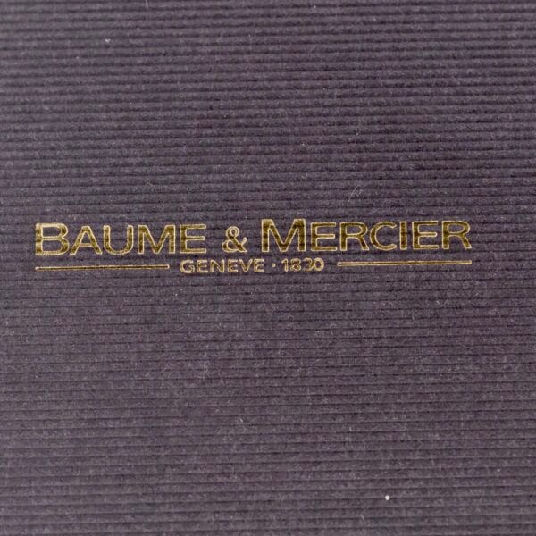 Close-up of a textured black surface with BAUME & MERCIER GENEVE 1830 embossed in gold, echoing the elegance of the Baume Mercier 18k Gold Calendar.