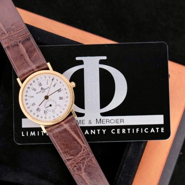 A Baume Mercier 18k Gold Calendar watch with a brown leather strap sits on a black warranty card inside the box, showcasing its classic white dial and intricate numbering that highlight timeless elegance and craftsmanship.