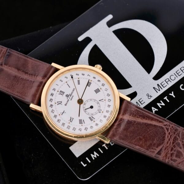 A Baume Mercier 18k Gold Calendar wristwatch with a white face, brown leather strap, Roman numerals, and a small seconds subdial rests on a black card with the logo.