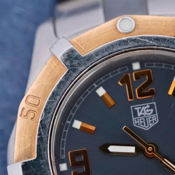 Close-up of a Tag Heuer 2000 Exclusive watch face with gold-toned numerals and hour markers on a grey background. It features an 18k gold-accented metallic bezel and prominent branding in the center, with minor scratches enhancing its authentic charm.