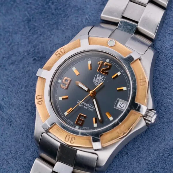 The Tag Heuer 2000 Exclusive 18k Gold Bezel wristwatch features a silver and gold design, water resistance up to 200 meters, a dark face with orange numerals on a blue textured background showing the date as 10, and a sophisticated metal link bracelet.