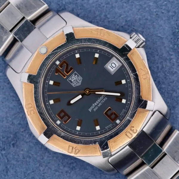 The Tag Heuer 2000 Exclusive wristwatch features an 18k gold bezel, silver metal bracelet, and a dark gray dial with large numerals at 6 and 12. It includes a date window at 3 oclock, set against a blue surface for an elegant, classic allure.