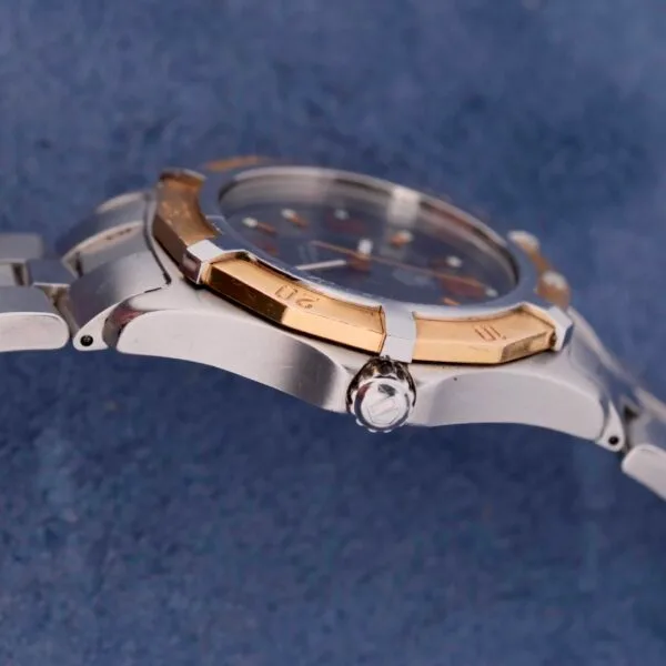 Close-up of a Tag Heuer 2000 Exclusive wristwatch with an 18k gold bezel and metallic band. The polished watch face is partially visible against a textured blue fabric background.