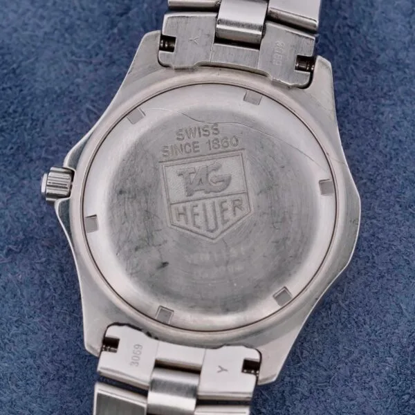 The back of a TAG Heuer 2000 Exclusive 18k Gold Bezel watch displays an engraved logo and Swiss Since 1860 on a brushed silver surface, elegantly resting on textured blue fabric.