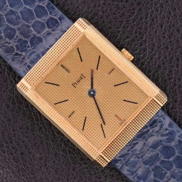 The Piaget Tank Ref 9151 18k Gold Manual Wind features a square gold watch with a textured dial, minimal black hour markers, and a blue leather strap. Its elegant design against a dark background makes it a timeless statement piece.