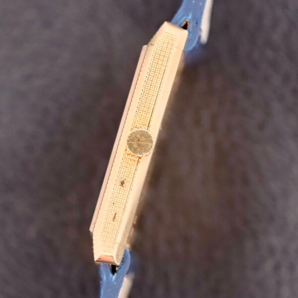 Close-up of the Piaget Tank Ref 9151 18k Gold Manual Wind wristwatch side, showcasing the textured case and small circular crown with a blue strap against a softly blurred background.