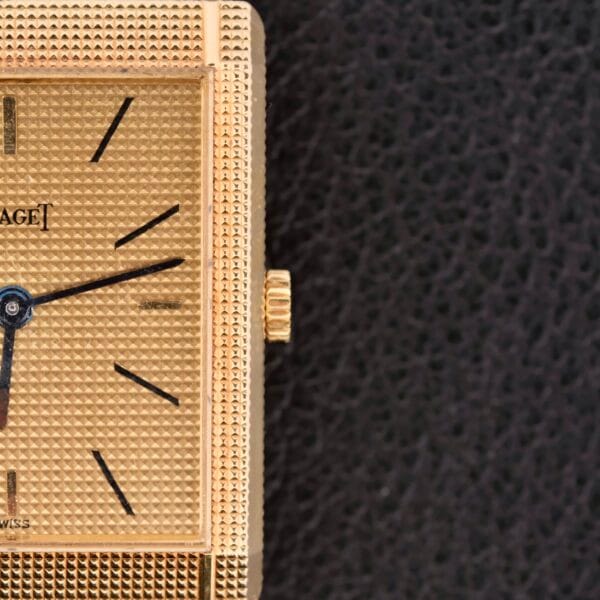 A close-up of the Piaget Tank Ref 9151, featuring a textured square face with minimalist black hour markers, is highlighted by a sleek gold crown against a refined black backdrop. This wristwatch is a manual wind model crafted from 18k gold.