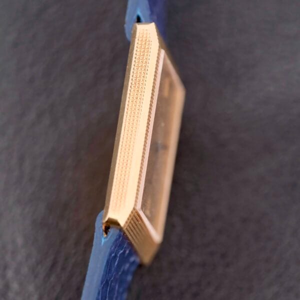 A close-up side view of the Piaget Tank Ref 9151 18k Gold Manual Wind watch highlights its rectangular gold-toned case with textured edges against a dark background. Featuring a blue band, it exudes elegance and timeless style.