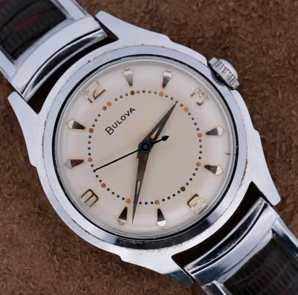 Close-up of a vintage 1956 Bulova Manual Wind Calibre 11AC wristwatch featuring a silver strap, cream-colored waffle dial, silver hour markers, numbers at 12, 3, 6, and 9 oclock, and two hands alongside a blued seconds hand on a textured brown background.