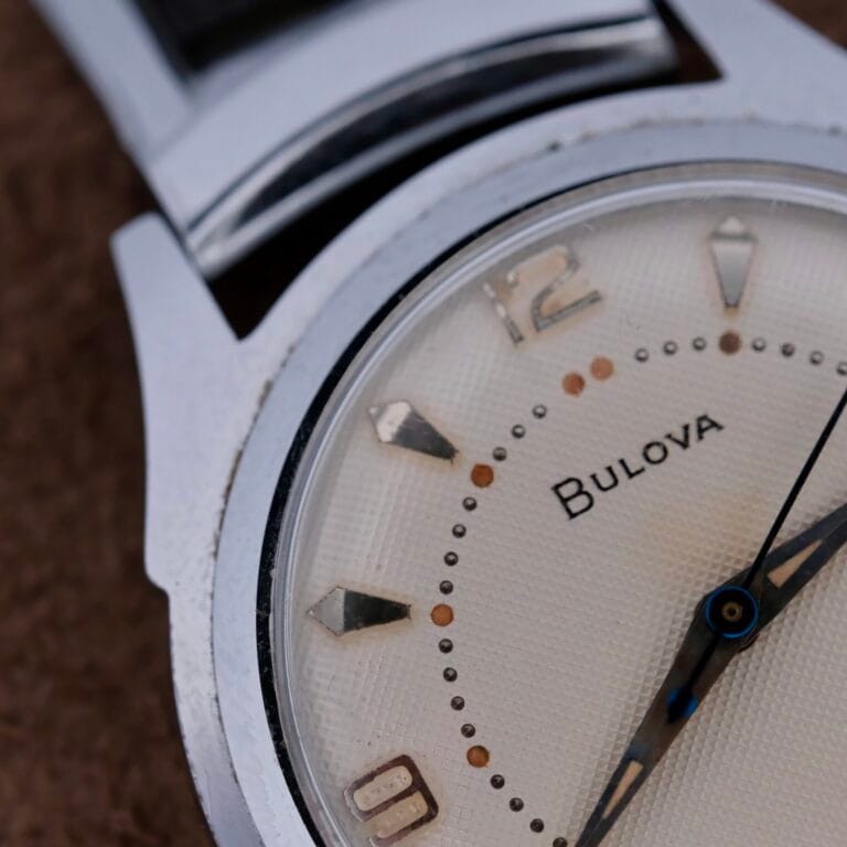 A close-up of the Bulova Manual Wind Calibre 11AC shows a beige waffle dial with silver markers and hands. It features a metallic bezel, black strap, and proudly displays Bulova on its dial.