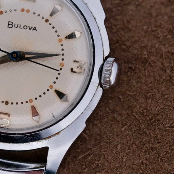 Close-up of a Bulova Manual Wind Calibre 11AC Waffle Dial wristwatch with a white textured dial, silver hour markers and hands. The partial view showcases the brand name and side crown against a textured brown background, echoing the style of the 1956 model.