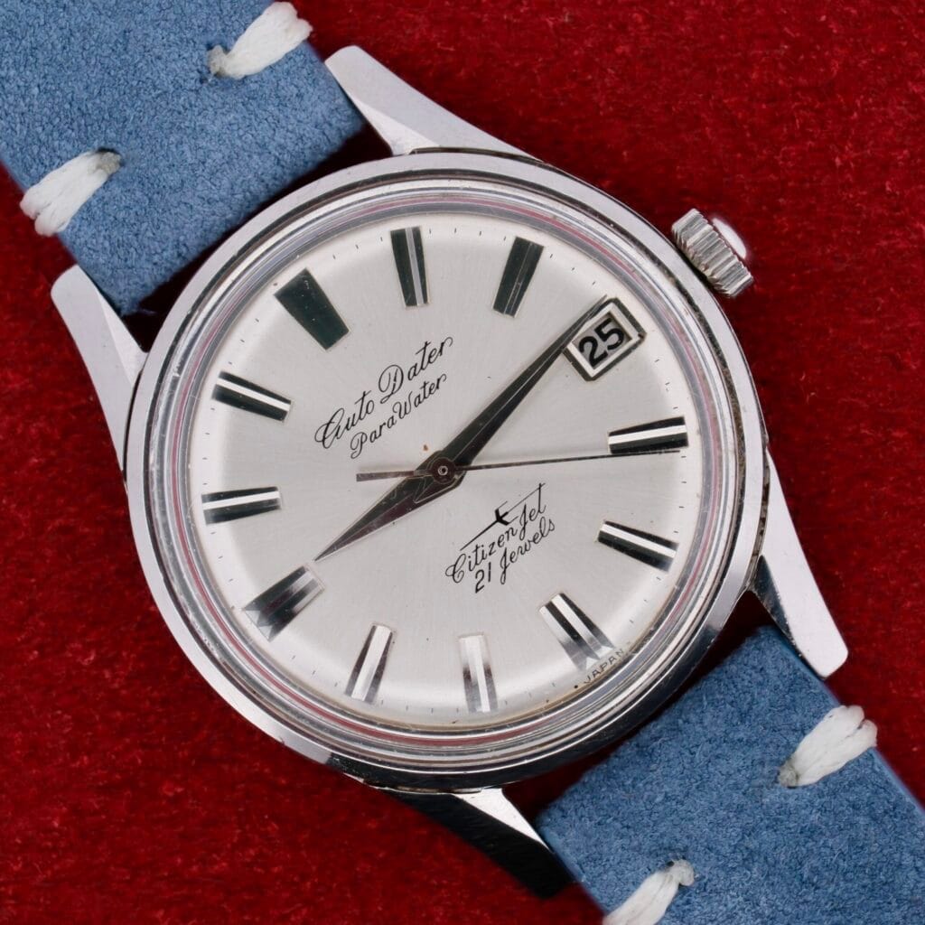 The Citizen Jet Auto Dater Automatic Para Water wristwatch features a silver dial with black hour markers, an auto date display set to the 25th, and includes a blue leather strap with white stitching. It is displayed on a red textured surface.